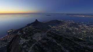 Buildings stand atop a rocky mountain overlooking an illuminated coastal city, harbor, and offshore island beneath a twilight sky.