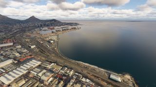 A coastal city surrounds a container port, with cranes, warehouses, and railway tracks lining the bay beneath mountains and scattered clouds.