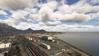A coastal harbor contains freight rail yards, warehouses, and container cranes, with a mountain-backed city beneath scattered cumulus clouds.