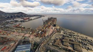 A coastal container port stretches beside calm water, with cargo cranes, docked ships, warehouses, and a hillside city beneath scattered clouds.
