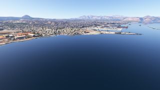 A coastal city borders blue water, with harbor cranes, docked ships, long breakwaters, and mountains beneath a clear sky.