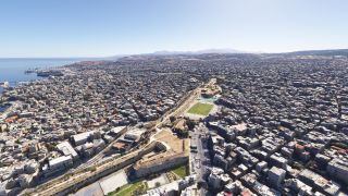 An aerial view shows a dense coastal city with stone fortifications, a green sports field, harbor docks, and distant hills.