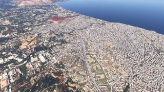 An aerial view shows dense coastal buildings, a curving highway, agricultural plots, and a shoreline stadium beside blue water.