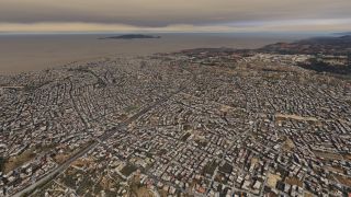 A dense coastal city stretches around a curving highway, with a harbor and offshore island beneath an overcast sky.