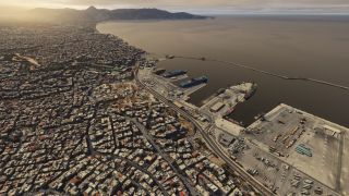 A dense coastal city borders a harbor with docked ships, container yards, and a long breakwater beneath distant mountains.