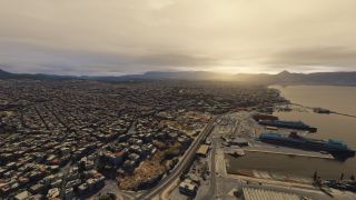 A dense coastal city borders a harbor with docked ferries, waterfront roads, and distant mountains beneath a cloudy, golden sky.