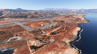 A coastal airport has a long runway, branching taxiways, and scattered buildings beside rocky shoreline, with a city and mountains beyond.
