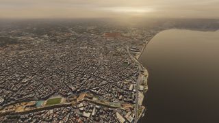 A dense coastal city borders a broad bay, with waterfront roads, an oval stadium, and fortified walls visible beneath hazy skies.