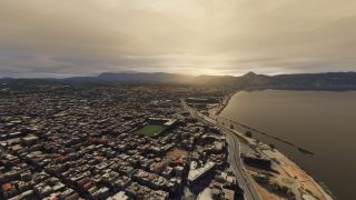 A dense coastal city borders a curved bay, with a waterfront road, green sports field, and mountains beneath golden clouds.