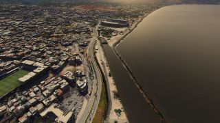 A coastal city spreads along a curving waterfront road, with two stadiums, dense buildings, and a long breakwater bordering the bay.