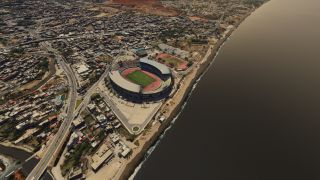 A coastal stadium contains a green playing field and red running track, with dense neighborhoods and curving roads bordering the shoreline.
