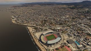 A coastal city stretches toward distant hills, with a waterfront stadium, red running track, swimming pool, and bridges in the foreground.