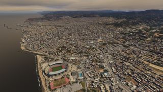 An aerial view shows a dense coastal city with a waterfront stadium, harbor breakwaters, winding highways, and hills beneath an overcast sky.