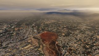 An aerial view shows dense buildings and winding roads surrounding a reddish plateau, with low clouds obscuring distant mountain ridges.