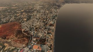 A coastal town stretches along a curved beach, with dense buildings, winding roads, swimming pools, and rocky hills inland.