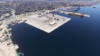 An aerial view overlooks a coastal harbor with docked ferries, cargo cranes, container yards, and dense urban buildings along the waterfront.