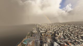A rainbow appears beneath thick clouds over a coastal city, with dense buildings, a waterfront road, and a curved seaside plaza.
