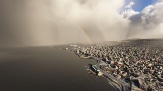 A rainbow appears beneath heavy clouds above a dense coastal city, with a curving waterfront road and ships in the harbor.