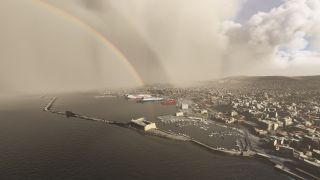 A rainbow arcs over a coastal city and harbor with docked ferries, a small-boat marina, and long stone breakwaters.
