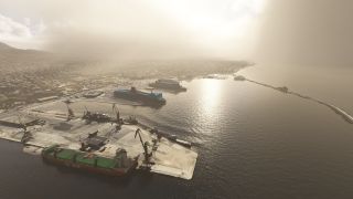 A coastal harbor contains docked ferries, a green-decked cargo ship, quay cranes, and a long breakwater beneath hazy sunlight.