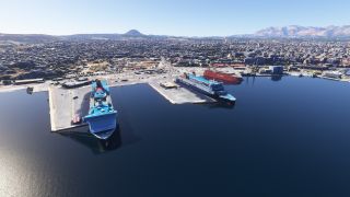 Large ferries sit moored at concrete harbor piers, with dense city buildings and distant mountains beneath a clear blue sky.