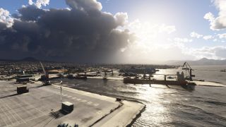 Cargo ships sit beside dockside cranes in a coastal harbor, with sunlit water, dense waterfront buildings, and dark clouds overhead.