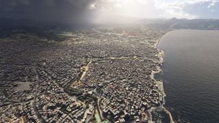 A dense coastal city borders a curving bay, with a small marina, inland mountains, and dark clouds contrasting with sunlit areas.