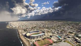 A large stadium with a running track sits beside a beach, with dense city buildings extending inland beneath towering clouds.
