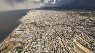 A dense coastal city stretches inland beneath dark clouds, with a waterfront stadium, running tracks, and curving highways visible from above.