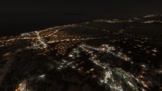 A nighttime aerial view shows a coastal city with illuminated streets, large industrial buildings, and dark water beyond the shoreline.