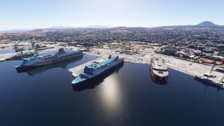 Three large ferries sit alongside harbor quays, with dockside cranes, a dense coastal city, and hills beyond.
