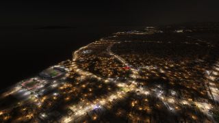 A coastal city glows at night with illuminated roads, dense neighborhoods, and a waterfront stadium beside an athletics track.