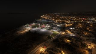 An aerial nighttime view shows a coastal stadium beside a floodlit running track, with illuminated streets extending through surrounding neighborhoods.
