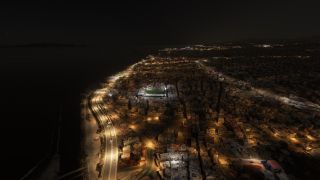 A coastal city stretches along dark water at night, with illuminated waterfront roads, dense buildings, and a floodlit stadium.