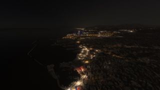 A coastal city stretches along a dark shoreline at night, with illuminated waterfront roads, boat-filled marinas, and angular harbor breakwaters.