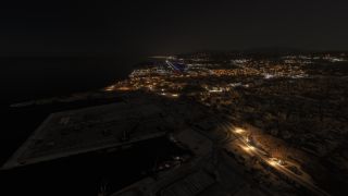 A coastal airport’s illuminated runway stretches beside dark water at night, with harbor docks, city buildings, and streetlights in the foreground.