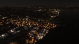 A coastal airport borders illuminated neighborhoods at night, with blue taxiway lights, parked aircraft, waterfront roads, and dark mountains beyond.