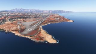 Runway 27 extends along a rocky coastline, with an aircraft near its threshold and urban development beneath distant mountains.