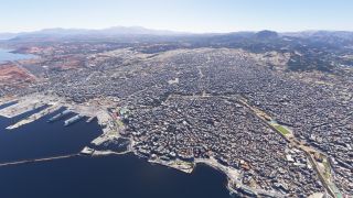 A dense coastal city surrounds a harbor with docked ships, a breakwater, and a marina, while an airport borders distant hills.