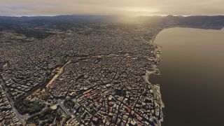 Dense city blocks line a curved waterfront, with winding roads, scattered green spaces, and mountains beneath a golden sky.