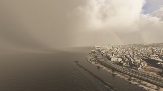 A coastal city borders dark water beneath heavy clouds, with a faint rainbow above waterfront roads and a long rocky breakwater.