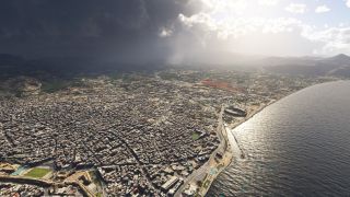 A dense coastal city stretches along a curving shoreline, with waterfront stadiums, inland mountains, and dark clouds contrasting with sunlit water.
