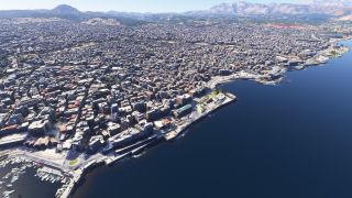 A dense coastal city stretches along a curved waterfront, with a boat-filled marina, shoreline roads, and mountains rising inland.