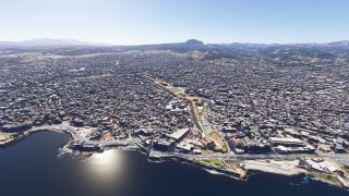 A dense coastal city stretches inland toward low mountains, with waterfront roads, a curved bay, and sports courts among tightly packed buildings.