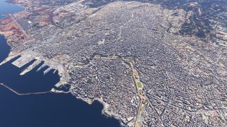A dense coastal city surrounds a harbor with docked ships and breakwaters, while an airport runway lies beside the shoreline.
