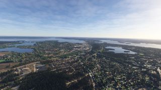 A lakeside city stretches across wooded terrain, with residential neighborhoods, industrial buildings, and numerous islands beneath a lightly clouded sky.