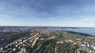 A city spreads across wooded terrain between broad waterways, with apartment blocks, winding roads, and distant islands beneath a lightly clouded sky.