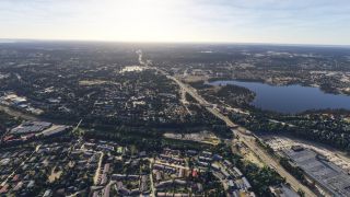 A broad highway runs through tree-lined residential neighborhoods beside a large lake, with commercial buildings and parking lots in the foreground.