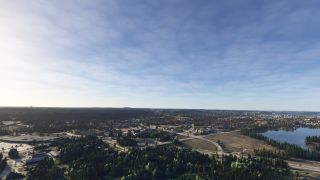 A town stretches across wooded terrain beside a lake, with intersecting roads, open fields, and low buildings beneath a lightly clouded sky.