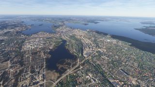 An aerial view shows dense neighborhoods, industrial buildings, and curving roads surrounding a small lake, with larger waterways and wooded islands beyond.